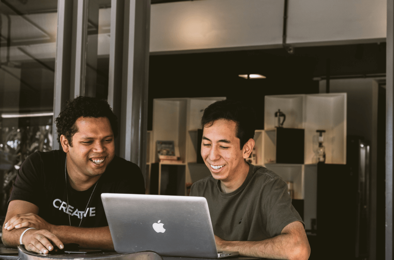 Two guys sits by a laptop laughing
