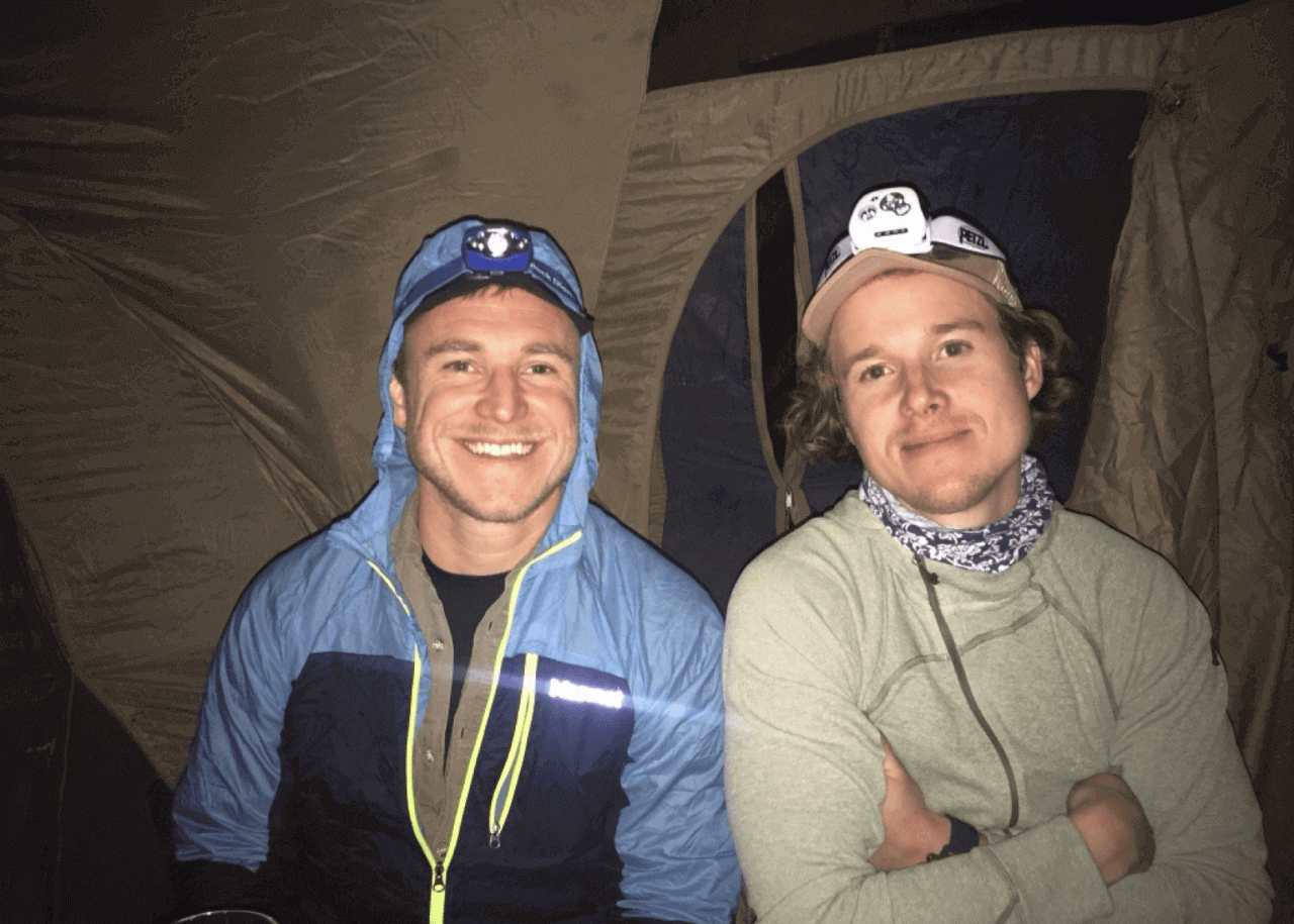 Two guys with headlights in front of a tent smiles towards the camera